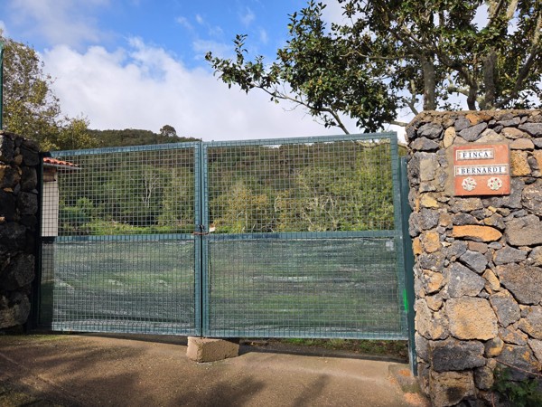 Entrada de la Finca Bernardi Entrada de una finca con gran portón metálico, muros de piedra volcánica y un cartel que dice Finca Bernardi, en un entorno rural y arbolado.