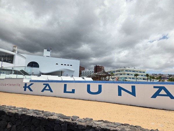 Vista exterior del club Kaluna en Tenerife, con muro blanco, letras azules, arena clara y un cielo cubierto de nubes oscuras.