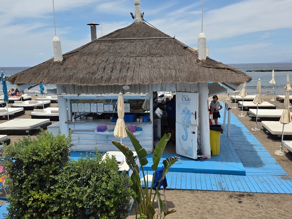 Chiringuito Le Club Playa Fañabé junto al mar, con techo de paja, pasarela azul, tumbonas blancas y playa al fondo en Tenerife.