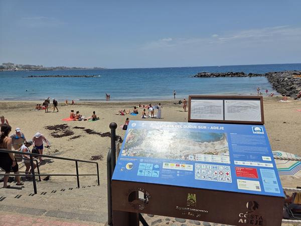 Vista de la Playa del Duque Sur en Adeje con cartel informativo en primer plano y turistas disfrutando del sol.
