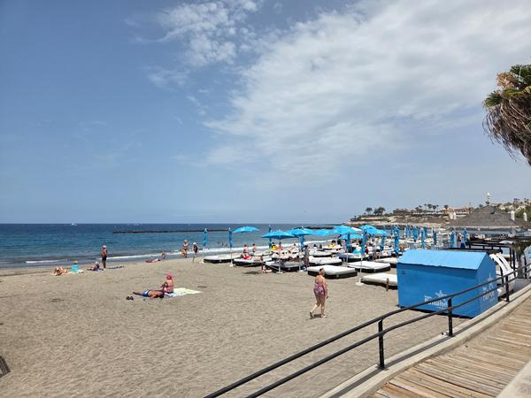 Playa Fañabé con sombrillas azules, tumbonas y paseo marítimo junto al océano en un día parcialmente nublado.