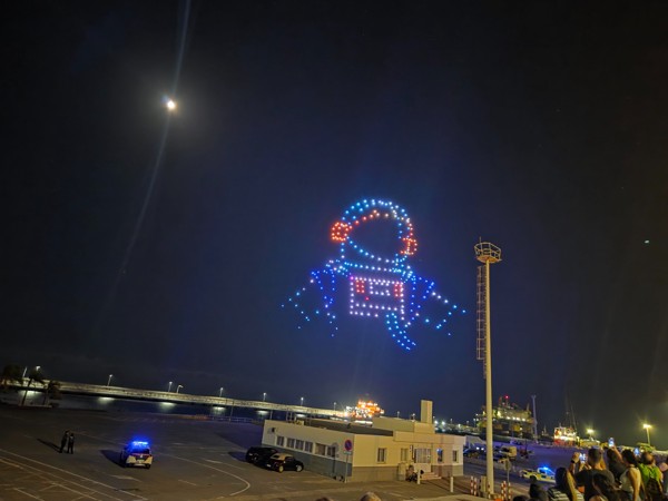 Drones en Plenilunio Espectáculo nocturno de drones en el cielo de Santa Cruz formando la figura de un astronauta, con la luna visible y el puerto al fondo.