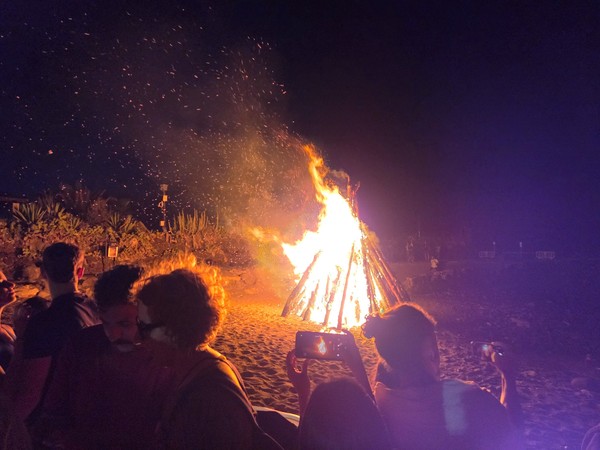 Gran hoguera encendida en la playa por la noche, rodeada de personas que observan y fotografían el fuego.