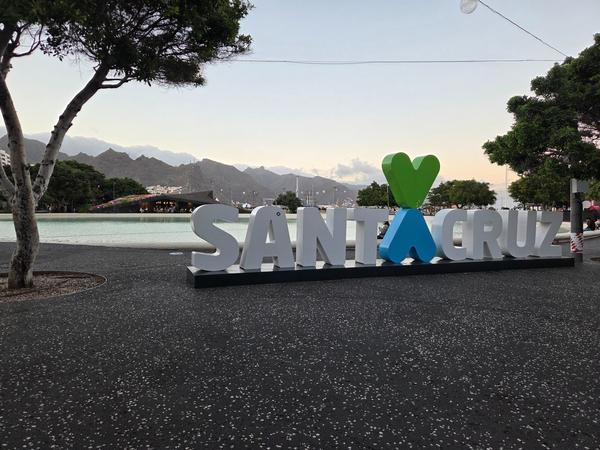 Letrero de Santa Cruz con símbolos de corazón verde y azul, y montañas al fondo al atardecer.